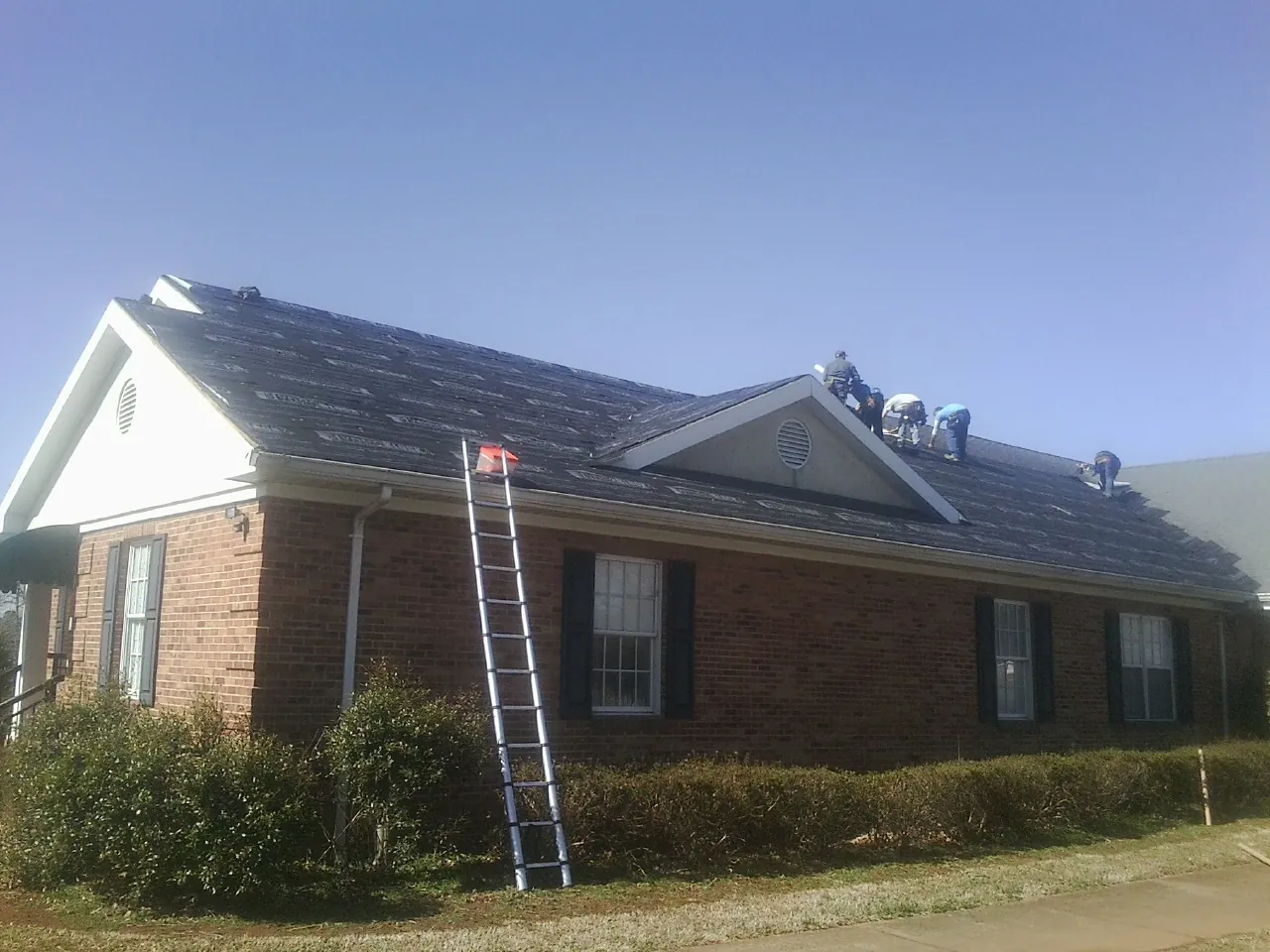 Crew tearing shingles off a brick ranch roof with a ladder set — Emergency Roof Repair work by our crew in Mc Alpin