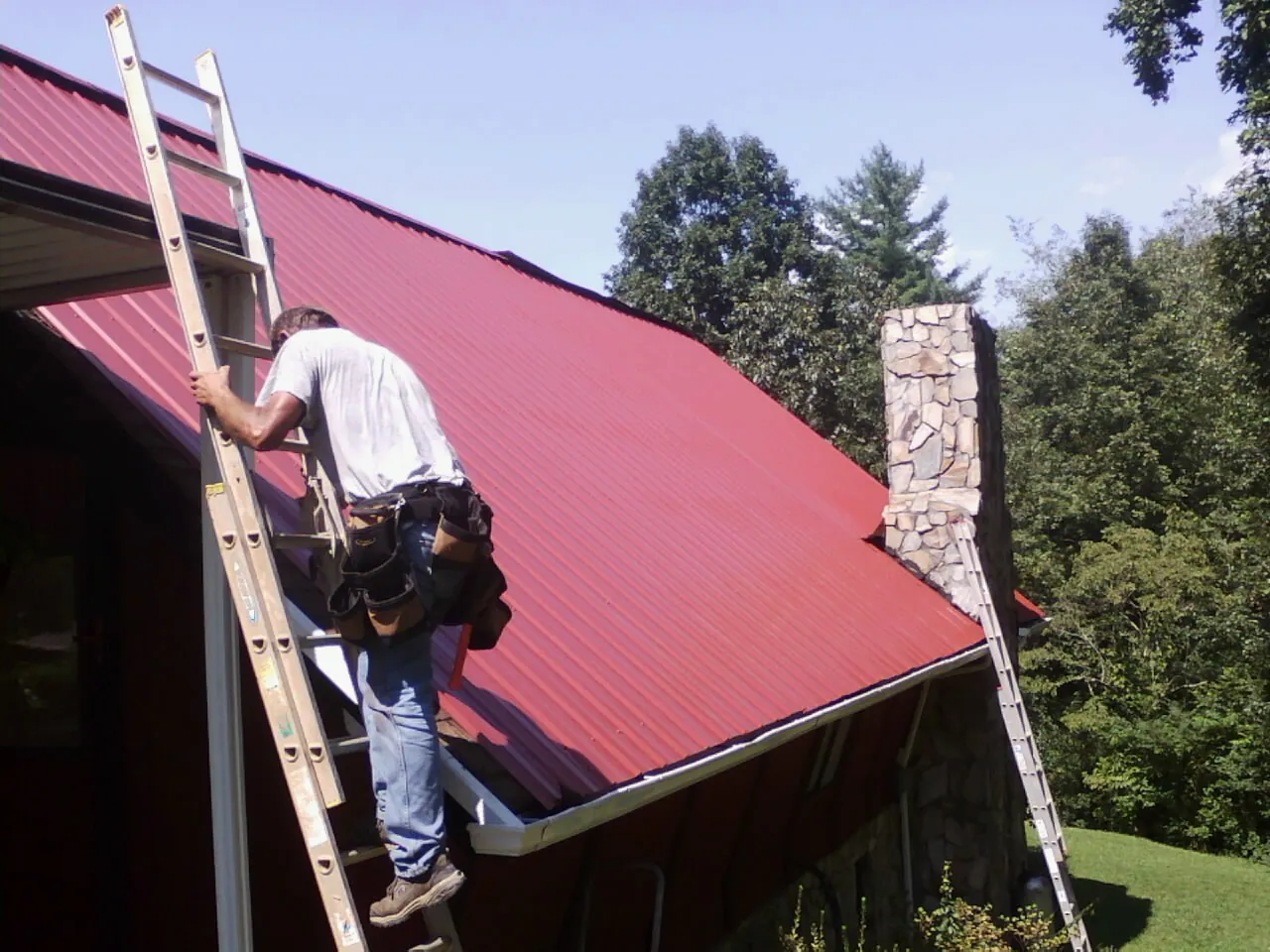Roofer on a ladder beside a red metal roof and stone chimney — Soffit & Fascia Repair by Roofing Brothers in Mc Alpin